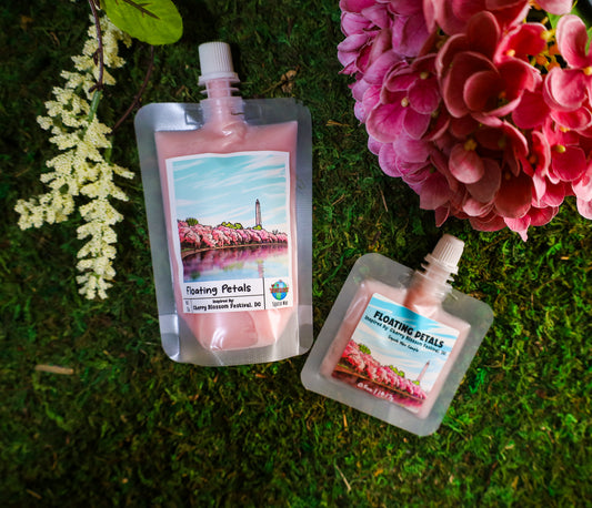 Two small pouches of 'Floating Petals' squeezable wax melt on grass with pink flowers in the background