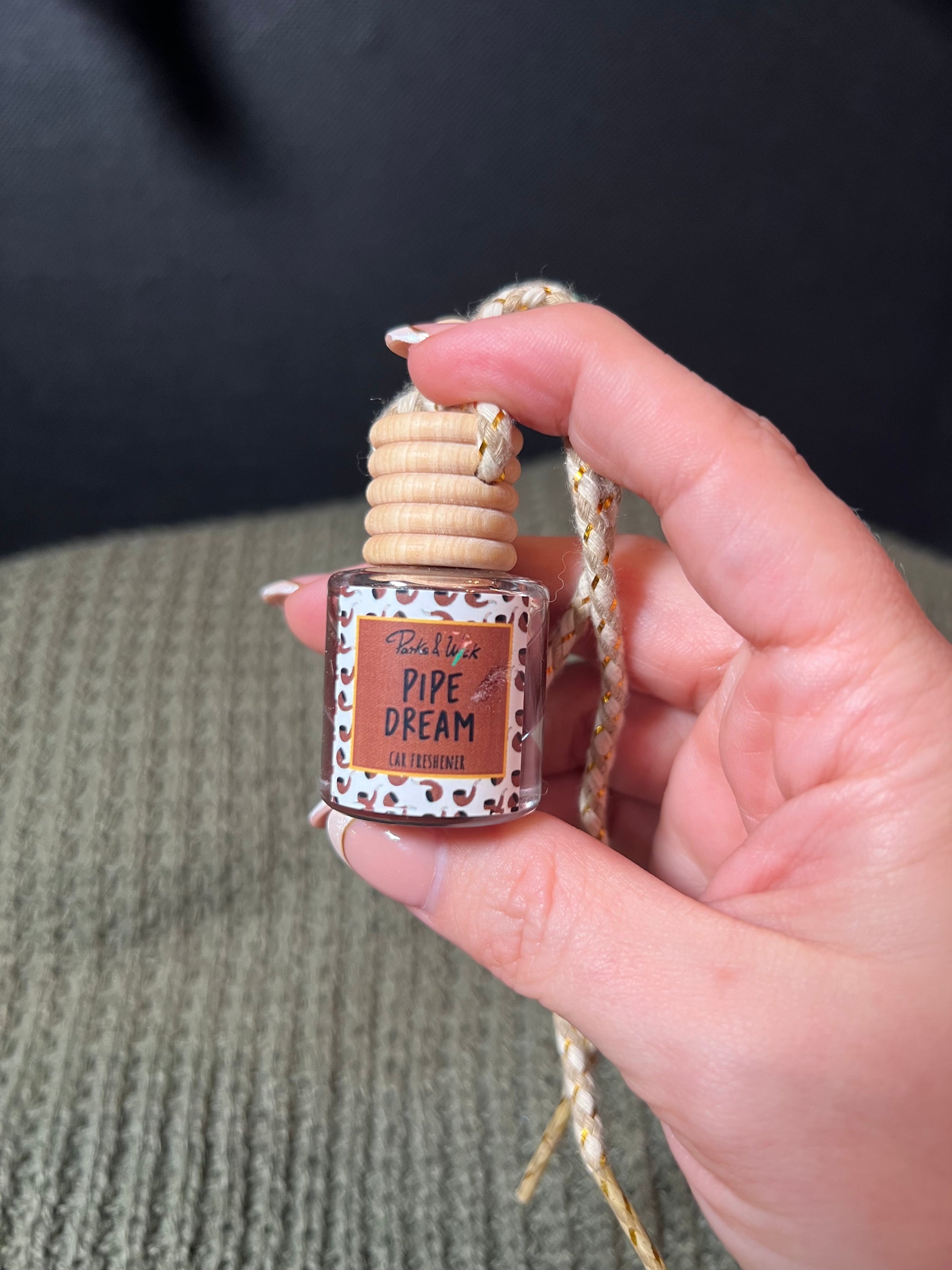 A hand holding a small brown car diffuser with text that reads 'PIPE DREAM' and a floral design, attached to a woven beige cord.