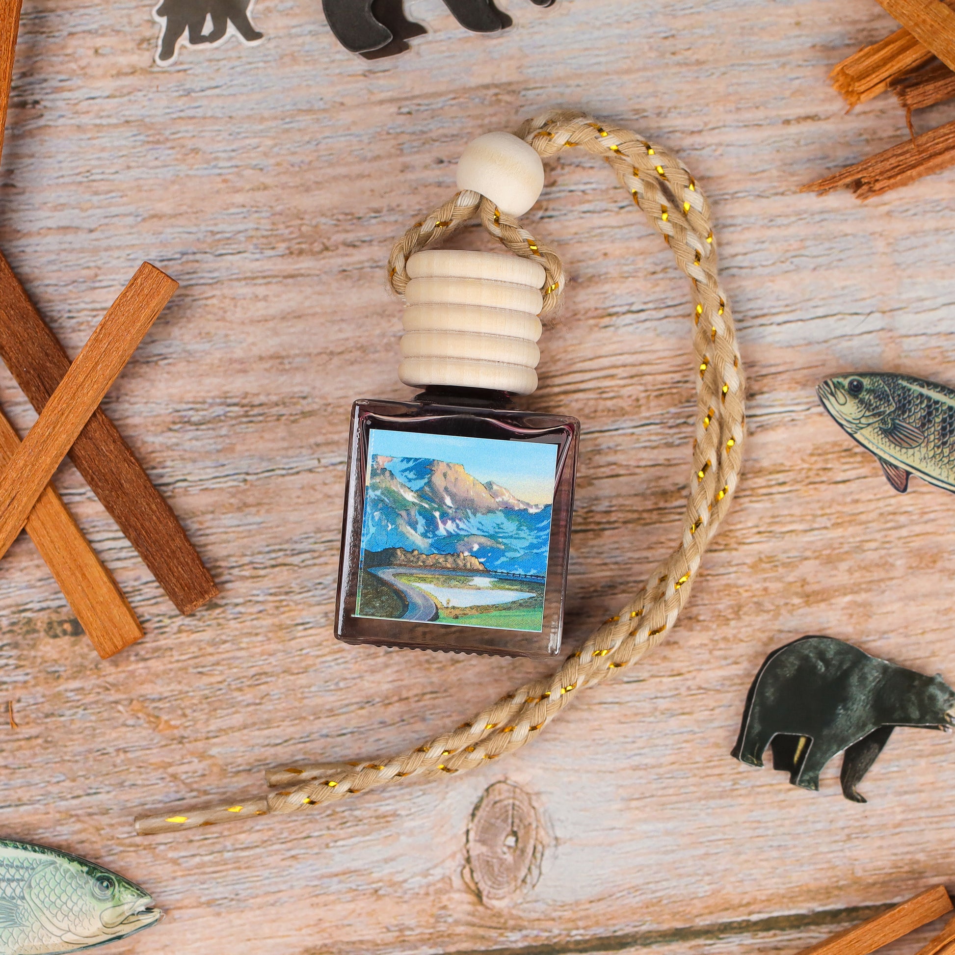 Artistic air freshener in a square glass bottle with a wooden cap and braided cord, featuring mountain scenery.